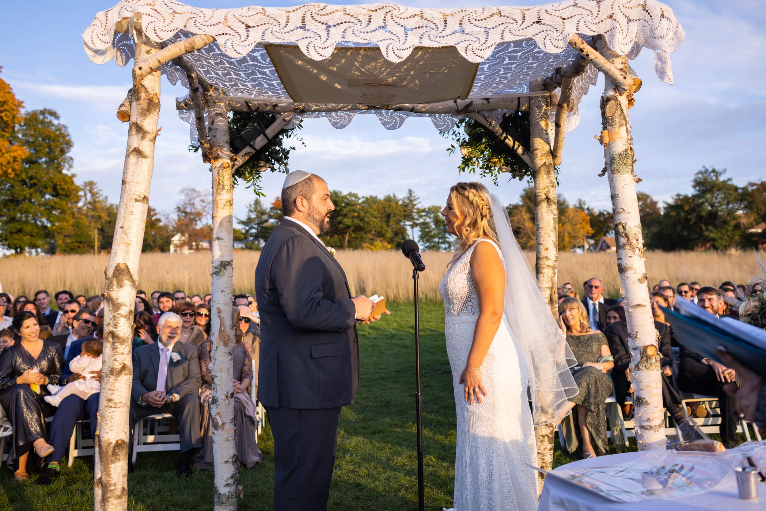 Bride and groom take their vows under a birch and lace chuppah while family and friends look on at sunset at fruitlands museum in Harvard ma