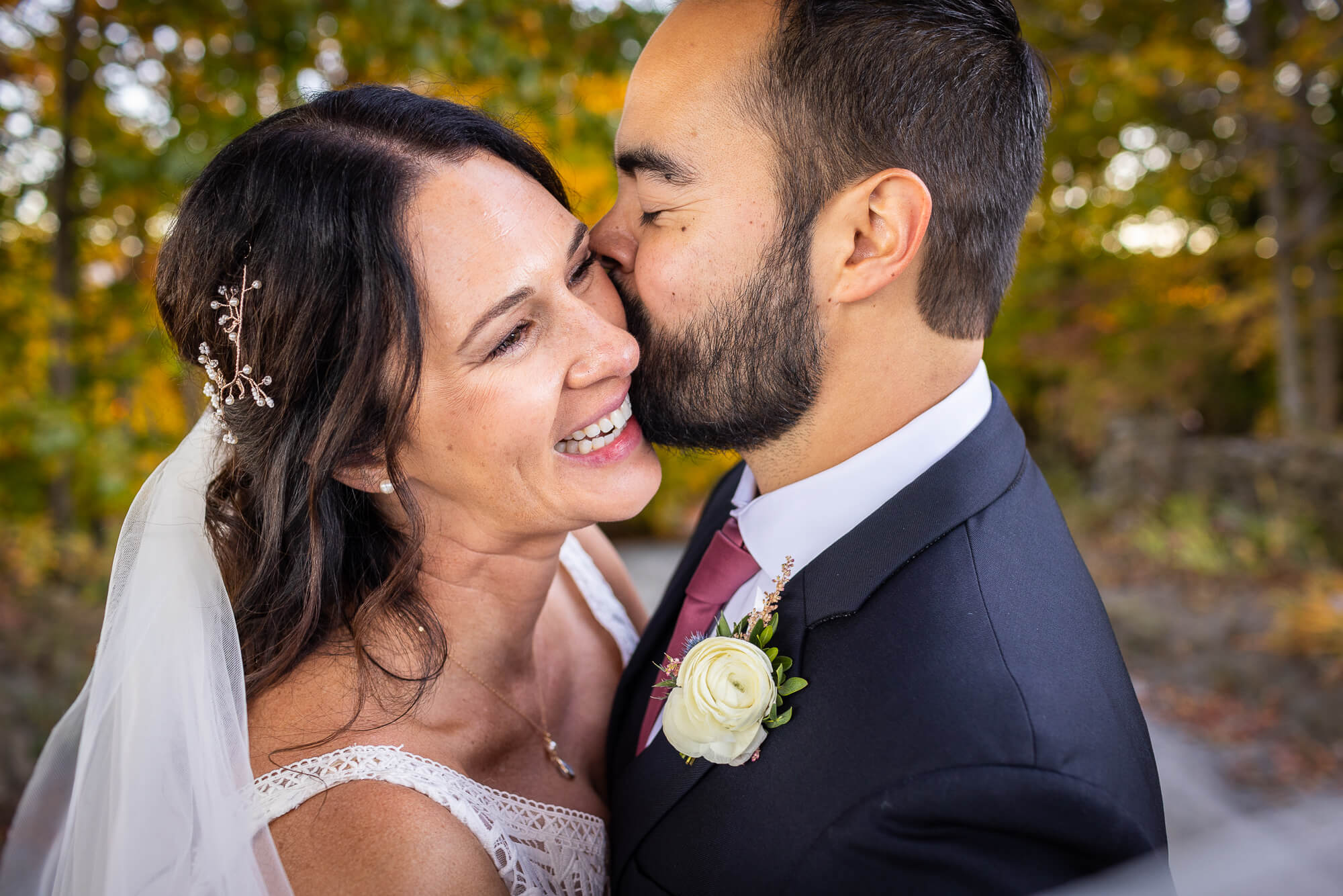 Groom kissing bride on the cheek she is smiling big in front of autumn leaves-Wedding-Photographer-audrey-cutler-photography