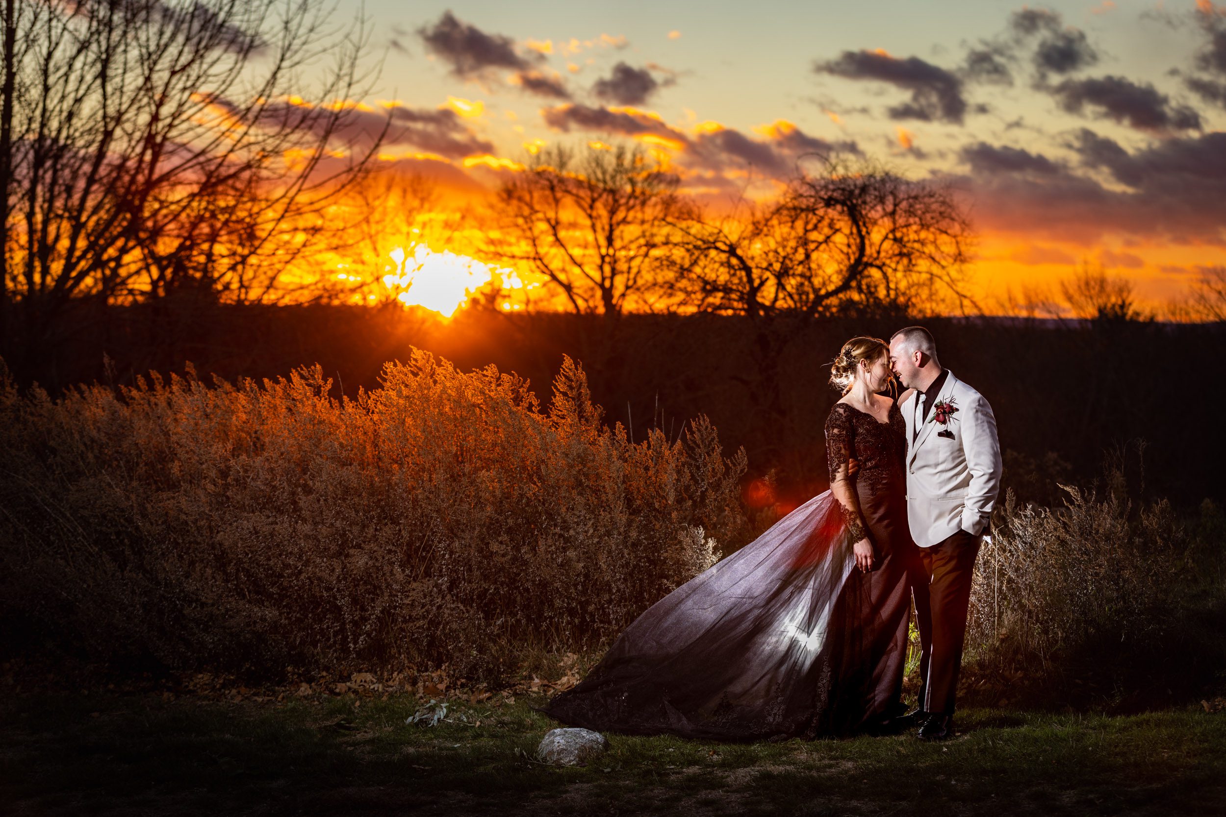 Bride in black wedding gown with groom in white tux who are well lit in front of a stunning orange sunset at Harrington farm in Princeton ma
