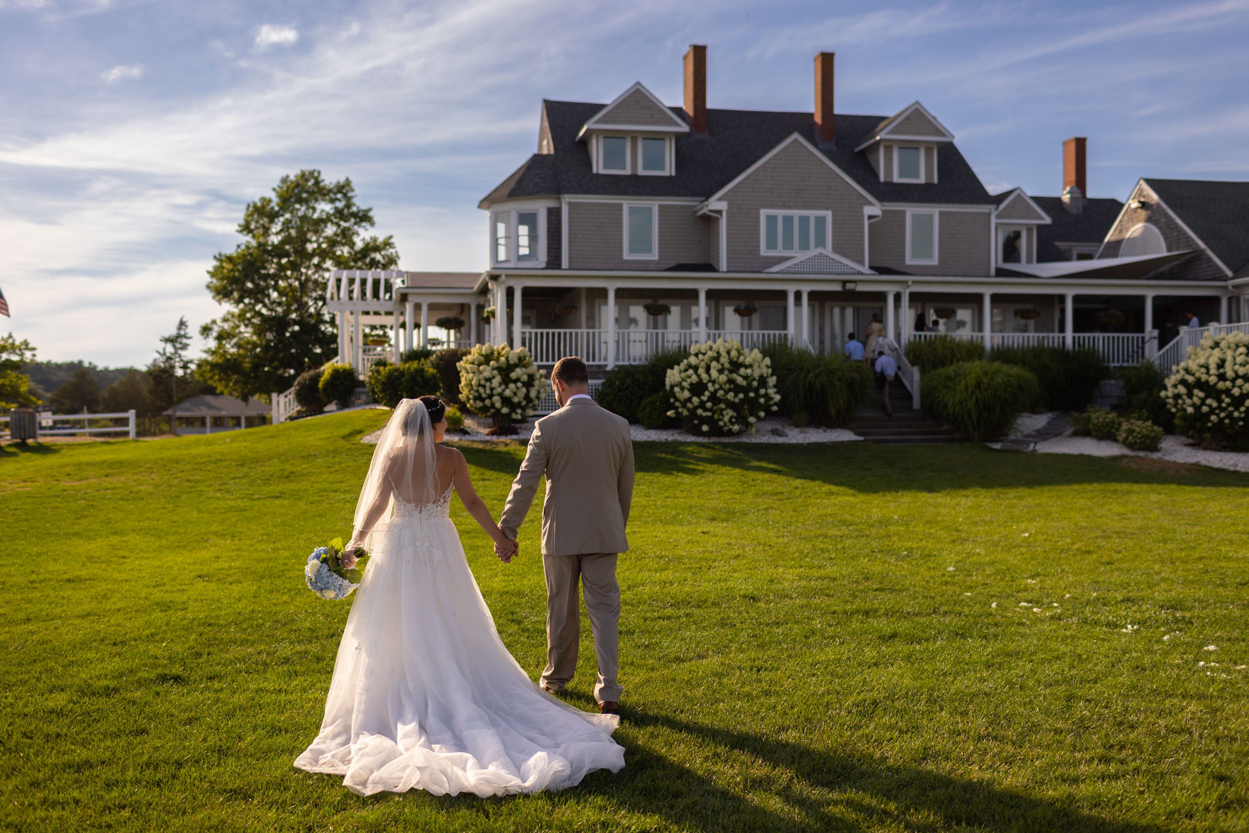 Bride and groom walk holding hand at sunset in front of estate at shining-tides-wedding