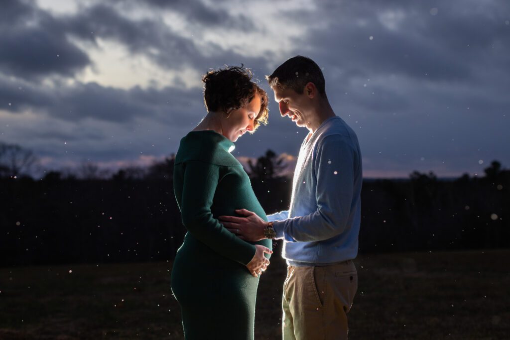 fall sunset snow maternity photography session at Harrington Farm in Princeton MA woman wearing green sweater dress holding baby bump with husband